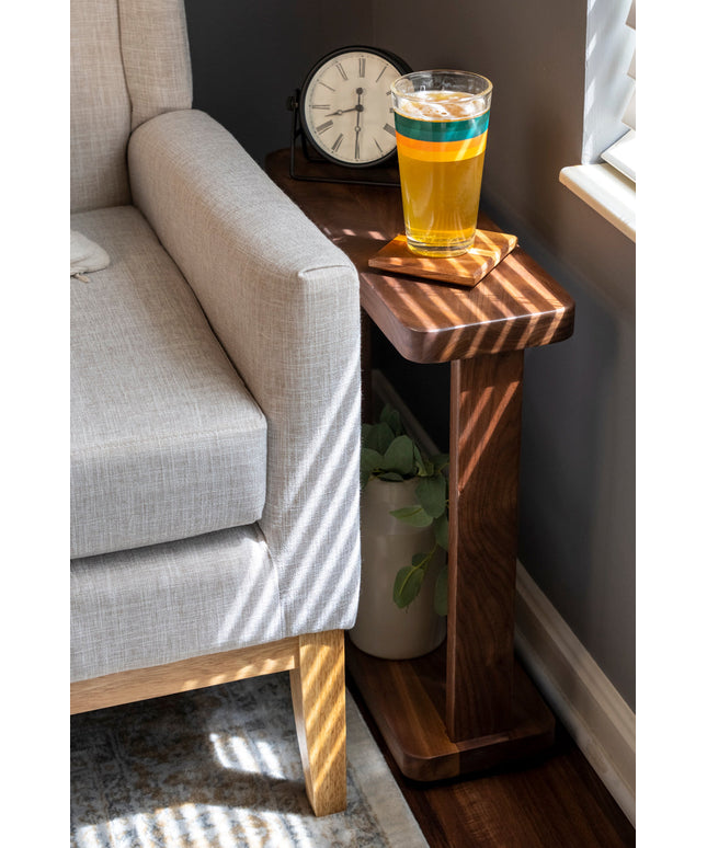 A small wooden side table with a glass of drink and a remote on top, placed between a couch and a wall.