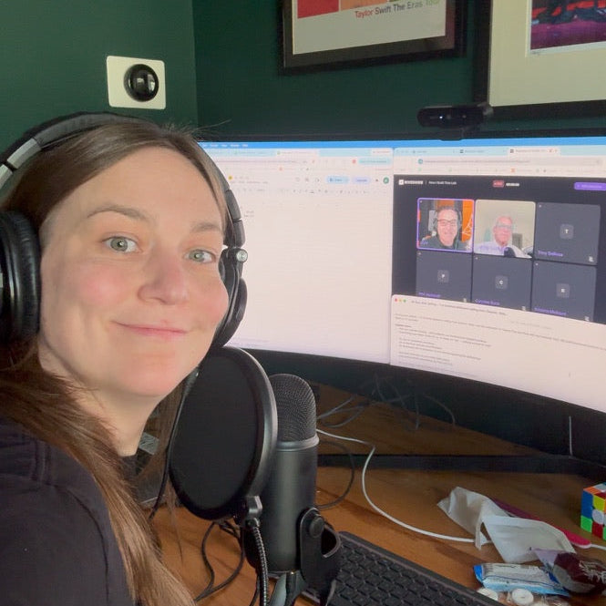 A woman wearing over-ear headphones sits at a desk with a curved computer monitor, microphone, keyboard, and office supplies in a home office setting.