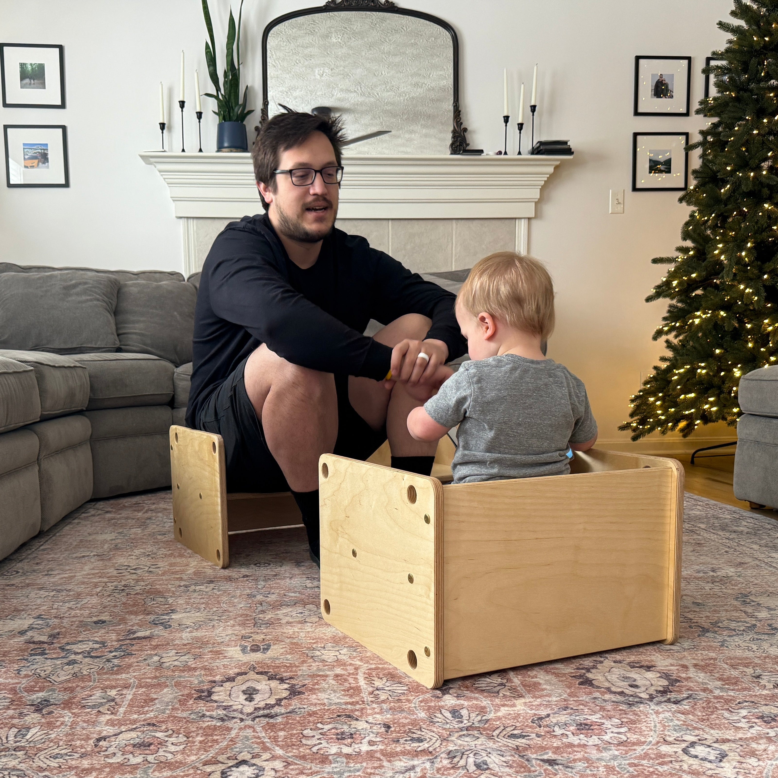 Dad and child sitting on wooden kids furniture in a living room decorated for Christmas.