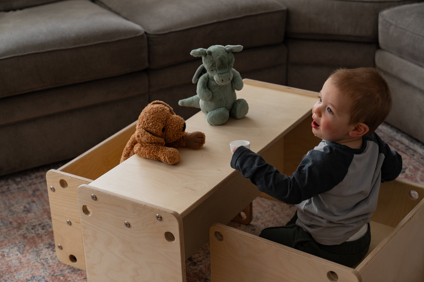 Child playing with stuffed animals on a wooden table in a living room.