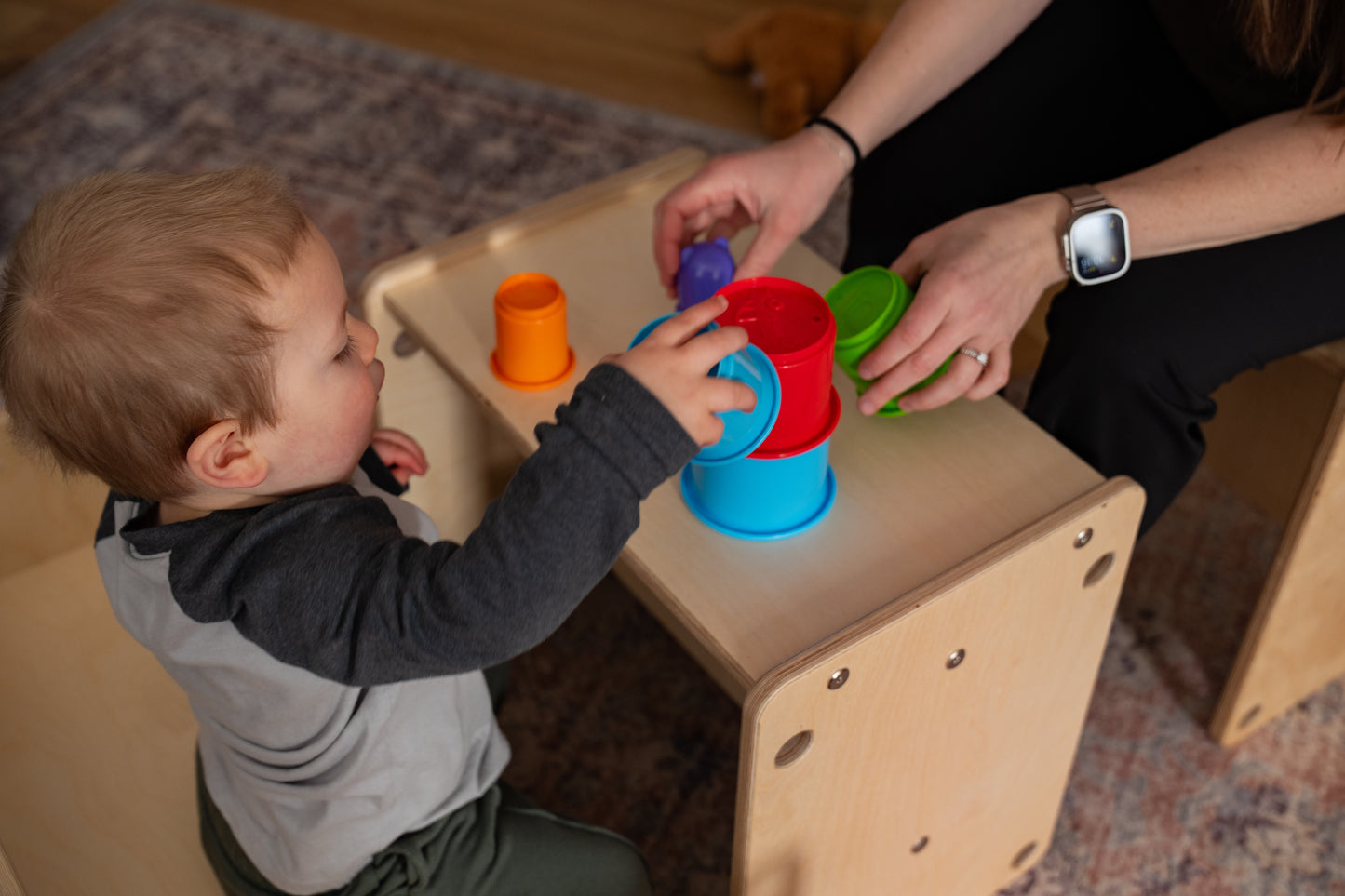 Child playing with colorful stacking cups on a wooden table with an adult's assistance.