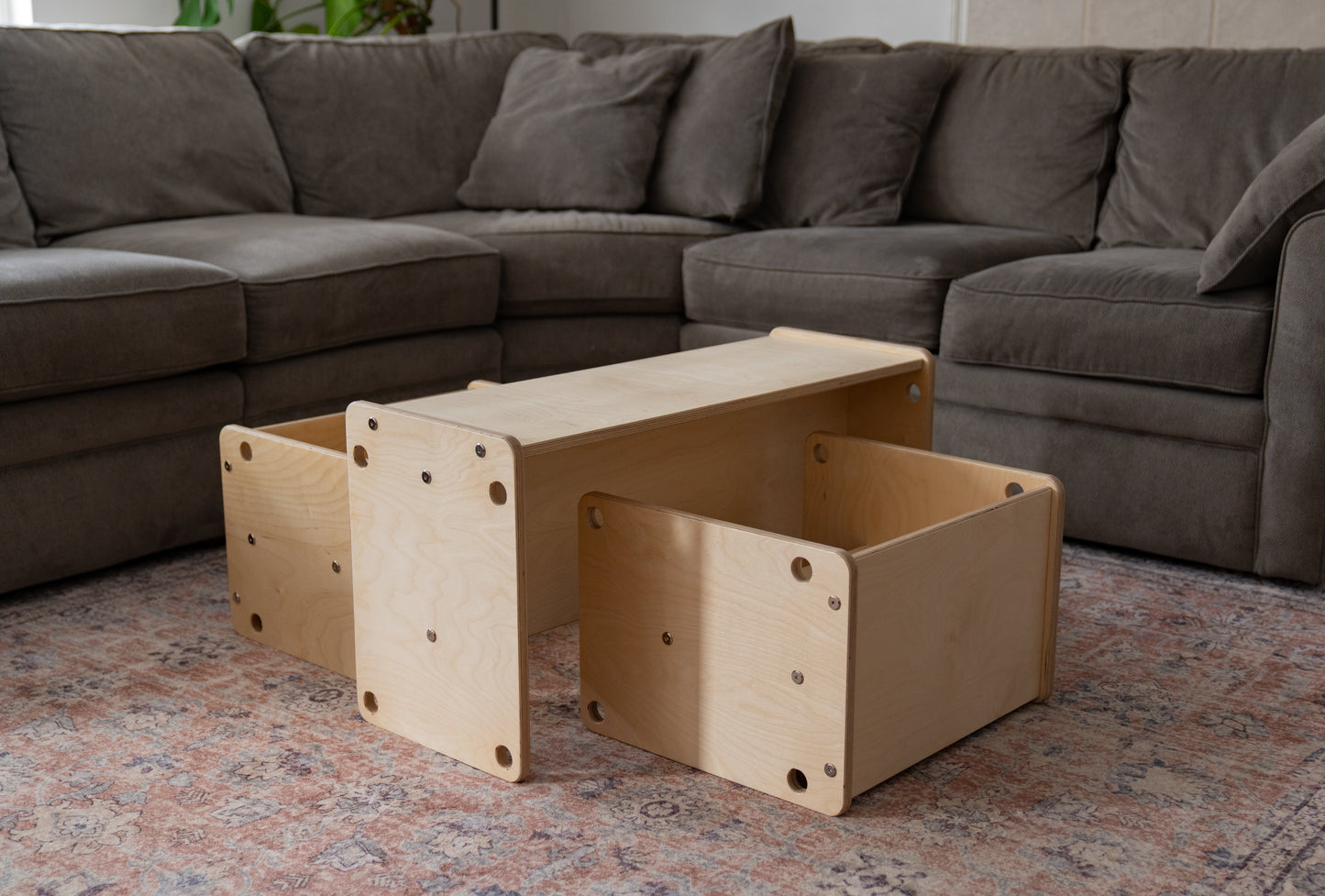 Wooden toddler table and chairs on a patterned rug with a gray sectional sofa in the background