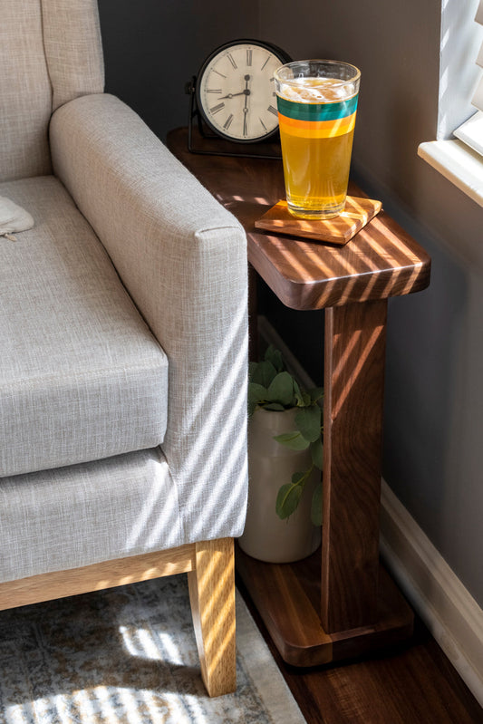 A small wooden side table with a glass of drink and a remote on top, placed between a couch and a wall.